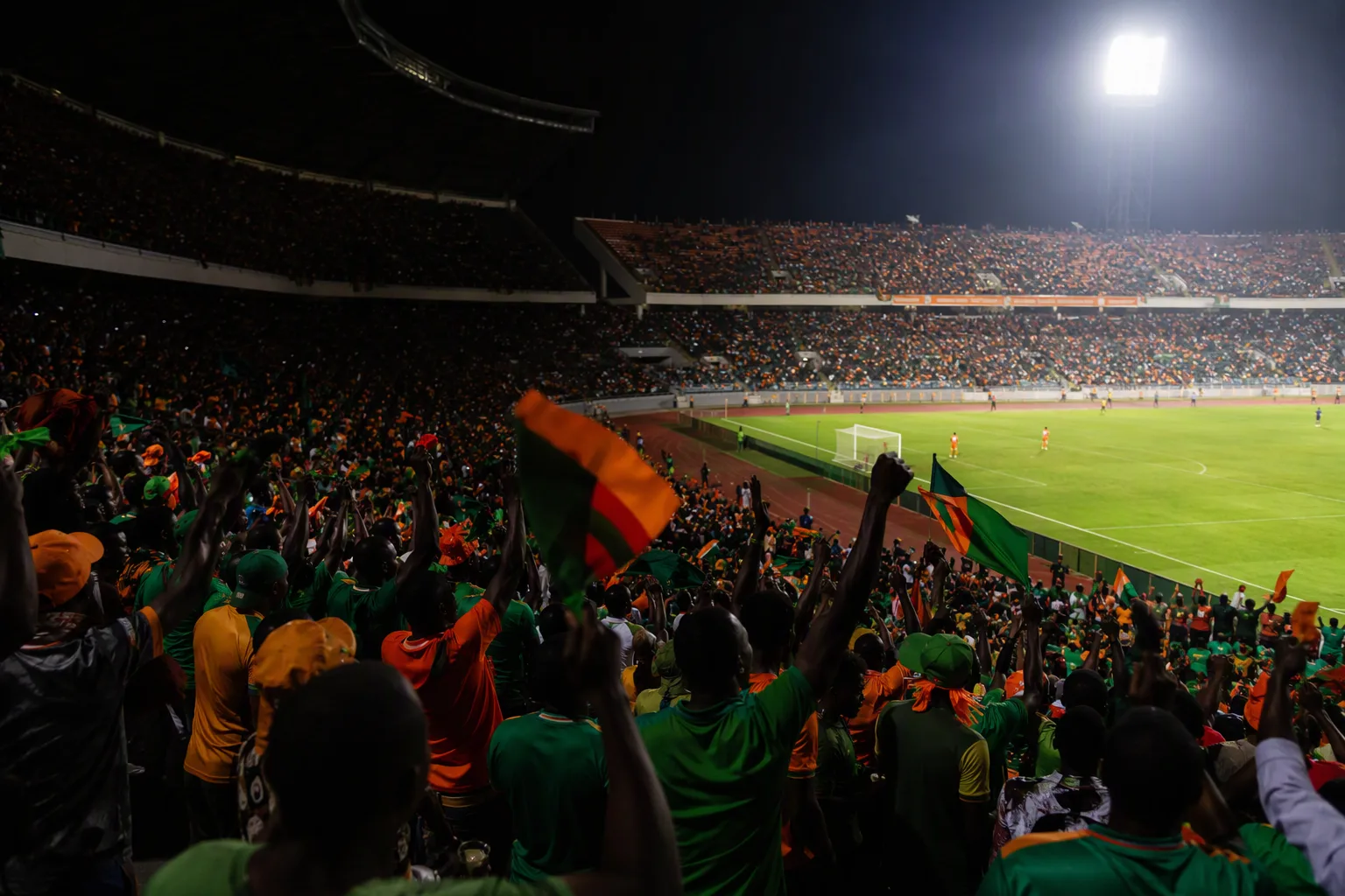 Zambian supporters celebrating under floodlights at a packed night match, one stand in darkness