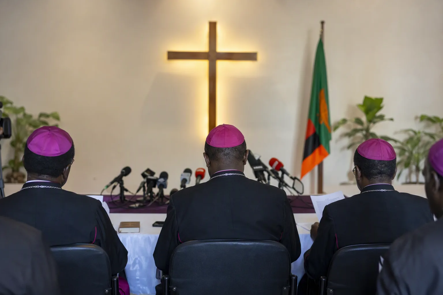 Catholic clergy seated at a long table before microphones at a Lusaka press briefing, seen from behind