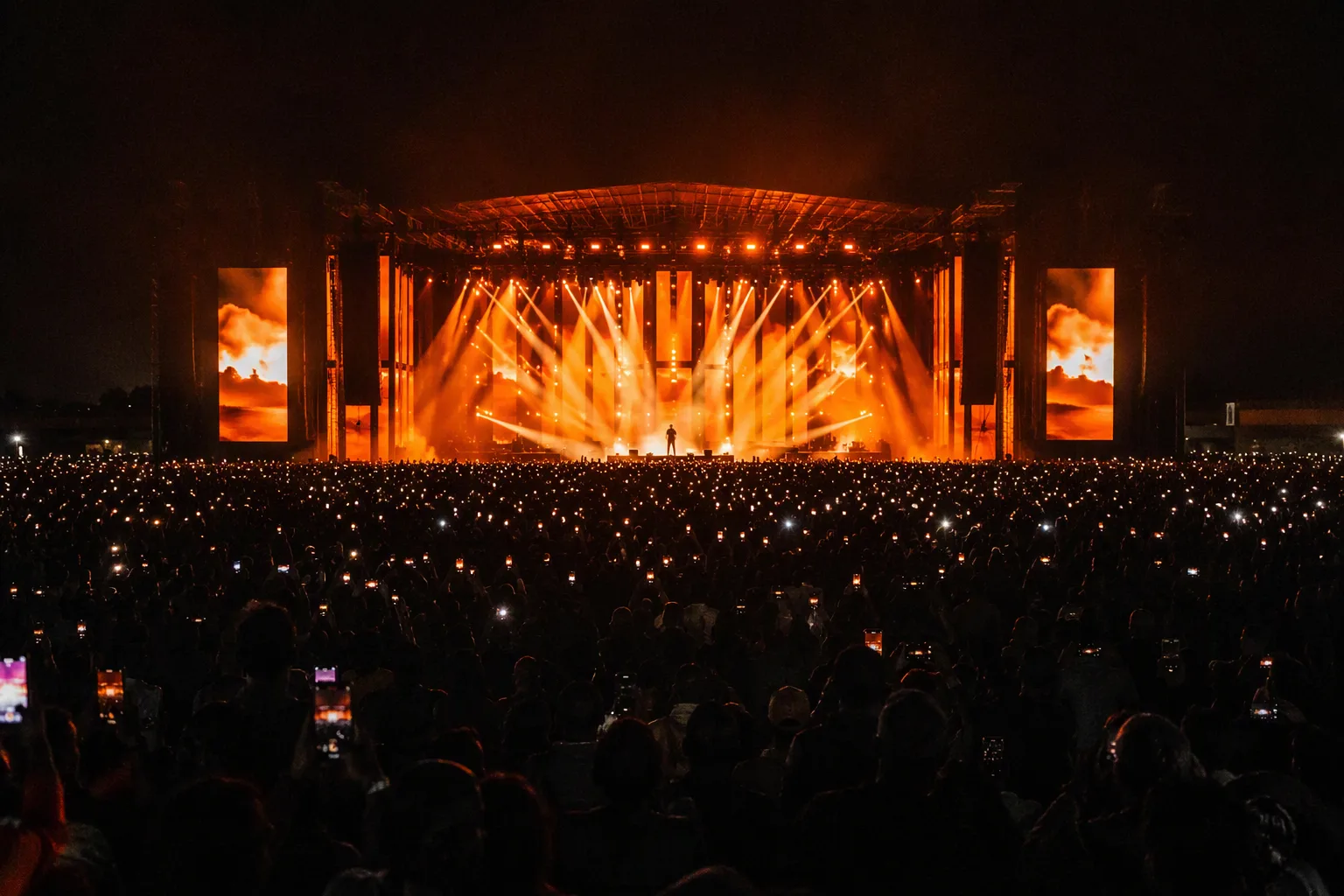 A massive outdoor concert stage at night bathed in copper and amber light, a sea of raised phone lights in the crowd