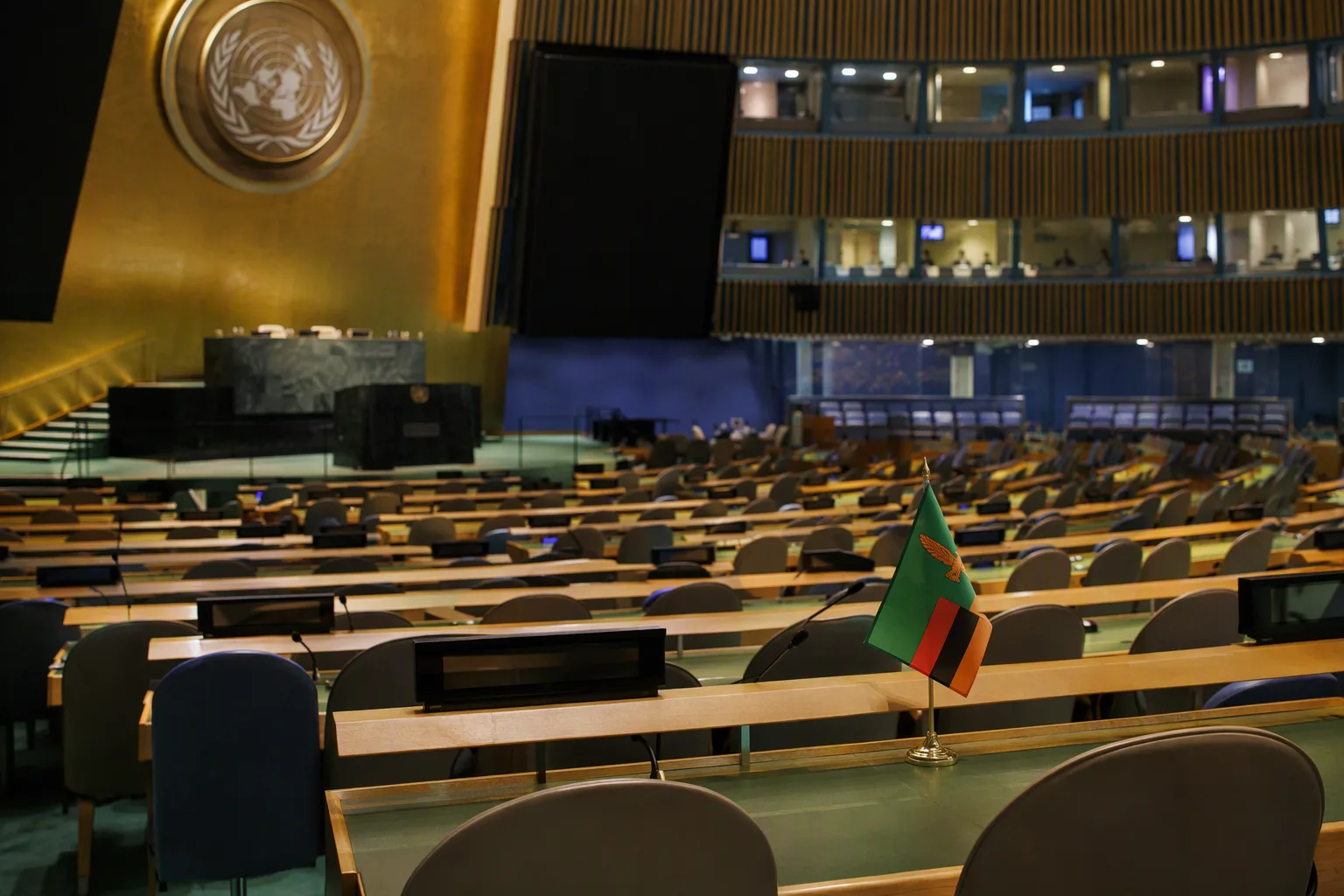 A grand UN-style assembly hall with curved rows of desks, a small Zambian flag on one delegation desk