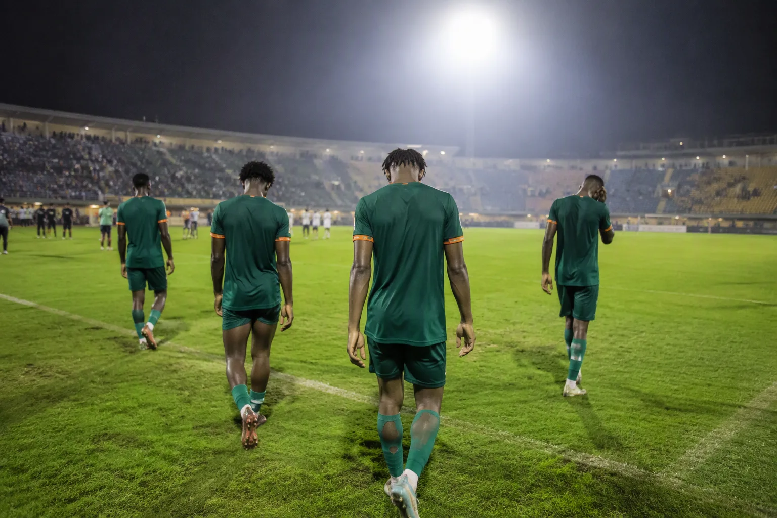 Football players in green shirts walking off a floodlit stadium pitch at night, dejected