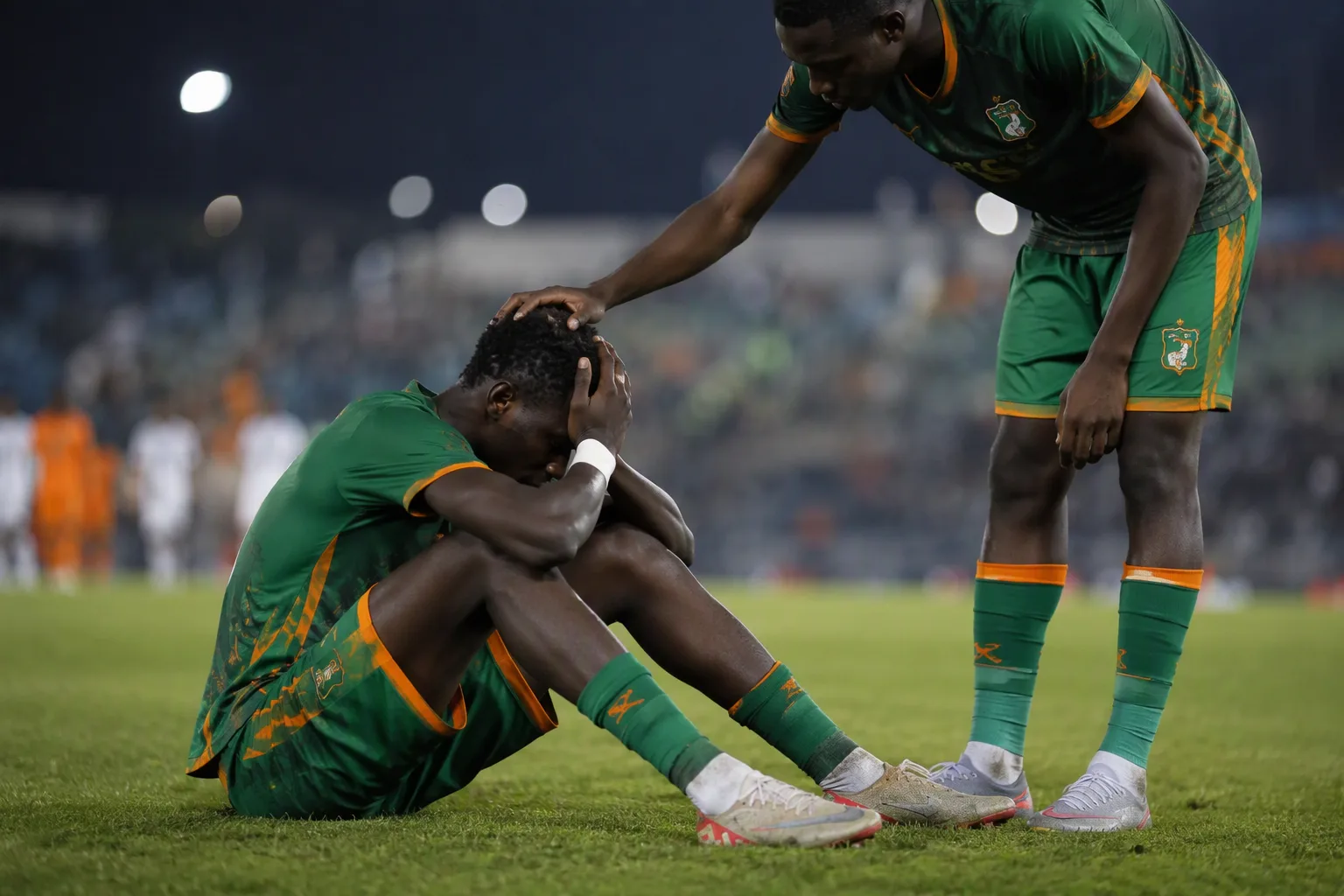 A young footballer in green and orange sitting on the pitch with his head in his hands after a defeat