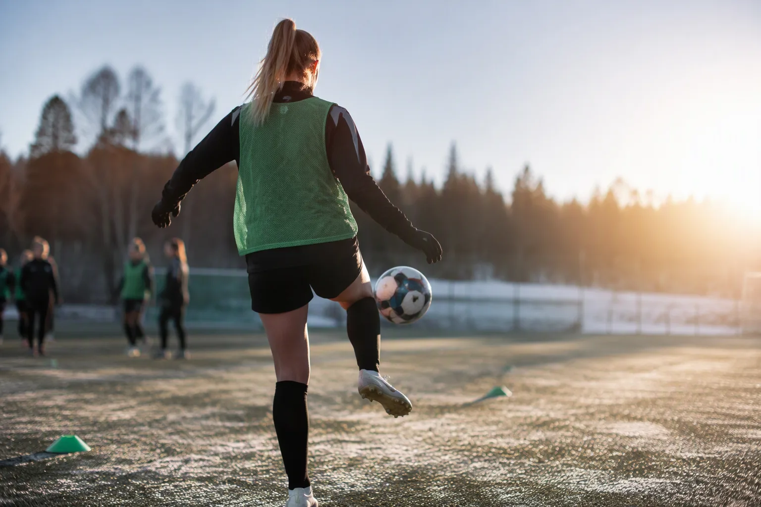 A young female footballer in a green training bib juggling a ball on a dewy pitch in pale morning light