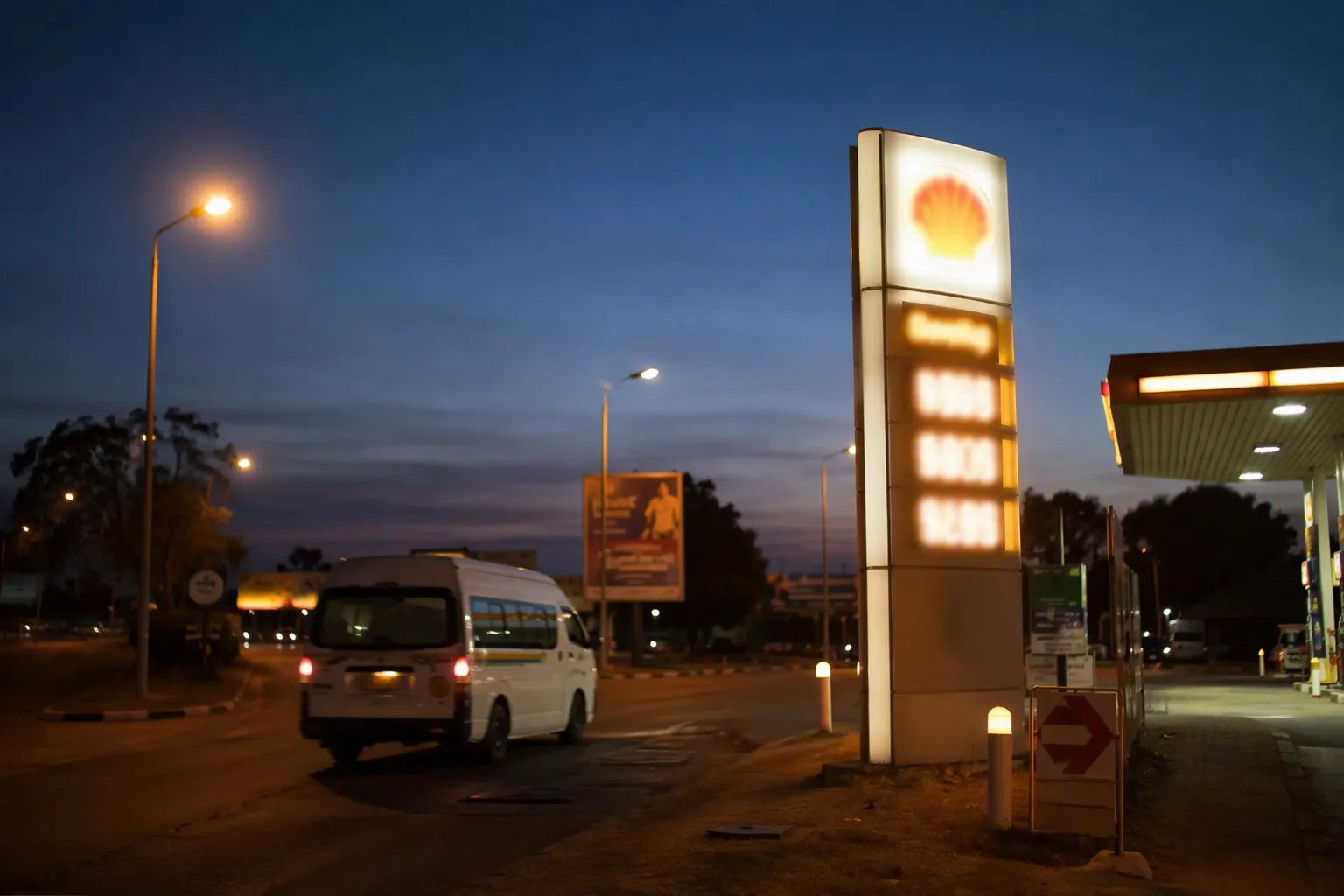 An illuminated fuel station price totem glowing at dusk with a minibus taxi passing